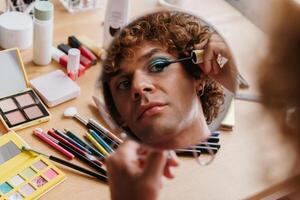 Top view of confident young gay man looking at his reflection in the mirror while applying make-up photo