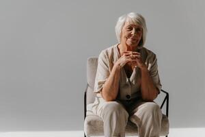 Relaxed gray hair senior woman sitting in comfortable chair and looking at camera in sunny studio photo