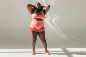 Full length of confident African plus size woman in sportswear adjusting hair while standing in fitness studio photo