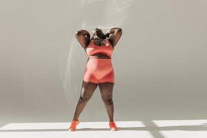 Full length of confident African plus size woman in sportswear adjusting hair while standing in fitness studio photo
