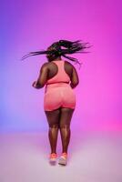Rear view of African curvy woman in sportswear dancing and looking confident against vibrant background photo