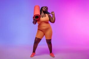 Happy African plus size woman in sportswear carrying exercise mat and smiling on vibrant background photo