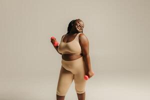 Confident plus size African woman in sportswear exercising with dumbbells on studio background photo