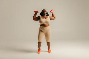 Full length joyful plus size African woman in sportswear exercising with dumbbells on studio background photo
