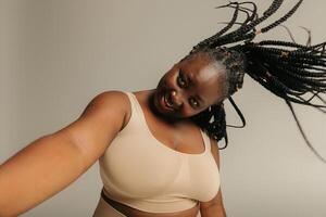 Joyful plus size African woman with dreadlocks making selfie while standing on studio background photo