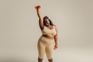 Confident plus size African woman in sportswear exercising with dumbbells on studio background photo