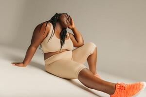 Young plus size African woman in sportswear looking relaxed while sitting against studio background photo