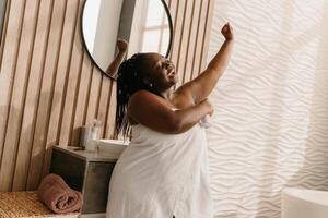 Happy plus size African woman covered in towel looking fresh and clean while spraying deodorant in bathroom photo