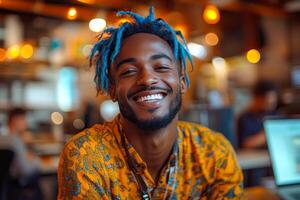 A man with blue hair and a yellow shirt is smiling. He is surrounded by people and there are two laptops in the background photo