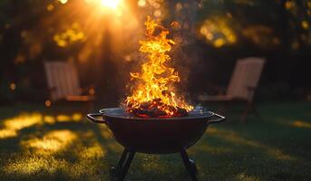 A fire is burning in a metal bowl on a grassy lawn. There are two chairs nearby, one on the left and one on the right. The fire is casting a warm glow on the scene, creating a cozy photo