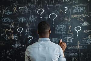 A man stands in front of a chaotic blackboard filled with question marks and scribbles, reflecting on his business strategy and the challenges ahead in his entrepreneurial journey to success photo