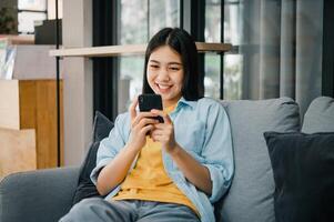 Smiling young asian woman using mobile phone checking social media,app playing game, shopping online, ordering delivery relax while sitting on a couch at home with laptop computer photo