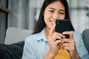 Smiling young asian woman using mobile phone checking social media,app playing game, shopping online, ordering delivery relax while sitting on a couch at home with laptop computer photo