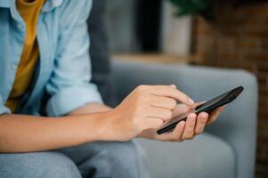 Smiling young asian woman using mobile phone checking social media,app playing game, shopping online, ordering delivery relax while sitting on a couch at home with laptop computer photo
