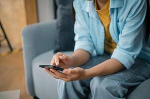 Smiling young asian woman using mobile phone checking social media,app playing game, shopping online, ordering delivery relax while sitting on a couch at home with laptop computer photo