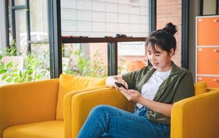 Smiling young asian woman using mobile phone checking social media,app playing game, shopping online, ordering delivery relax while sitting on a couch at cafe with laptop computer photo
