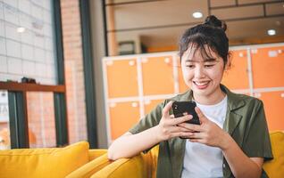Smiling young asian woman using mobile phone checking social media,app playing game, shopping online, ordering delivery relax while sitting on a couch at cafe with laptop computer photo