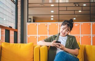 Smiling young asian woman using mobile phone checking social media,app playing game, shopping online, ordering delivery relax while sitting on a couch at cafe with laptop computer photo