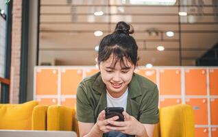 Smiling young asian woman using mobile phone checking social media,app playing game, shopping online, ordering delivery relax while sitting on a couch at cafe with laptop computer photo