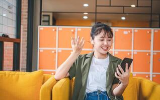 Smiling young asian woman using mobile phone checking social media,app playing game, shopping online, ordering delivery relax while sitting on a couch at cafe with laptop computer photo