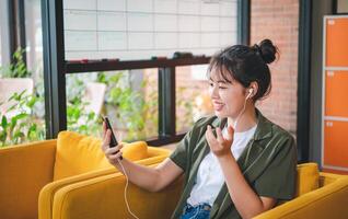 Smiling young asian woman using mobile phone checking social media,app playing game, shopping online, ordering delivery relax while sitting on a couch at cafe with laptop computer photo