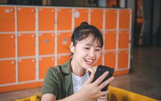Smiling young asian woman using mobile phone checking social media,app playing game, shopping online, ordering delivery relax while sitting on a couch at cafe with laptop computer photo