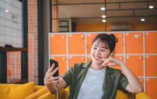 Smiling young asian woman using mobile phone checking social media,app playing game, shopping online, ordering delivery relax while sitting on a couch at cafe with laptop computer photo