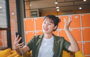 Smiling young asian woman using mobile phone checking social media,app playing game, shopping online, ordering delivery relax while sitting on a couch at cafe with laptop computer photo