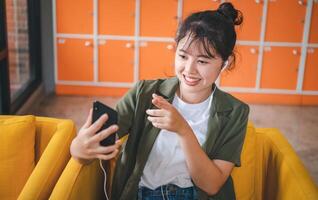 Smiling young asian woman using mobile phone checking social media,app playing game, shopping online, ordering delivery relax while sitting on a couch at cafe with laptop computer photo