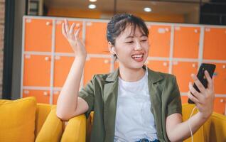 Smiling young asian woman using mobile phone checking social media,app playing game, shopping online, ordering delivery relax while sitting on a couch at cafe with laptop computer photo