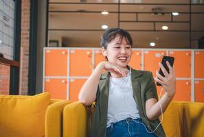 Smiling young asian woman using mobile phone checking social media,app playing game, shopping online, ordering delivery relax while sitting on a couch at cafe with laptop computer photo