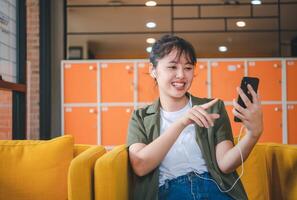 Smiling young asian woman using mobile phone checking social media,app playing game, shopping online, ordering delivery relax while sitting on a couch at cafe with laptop computer photo