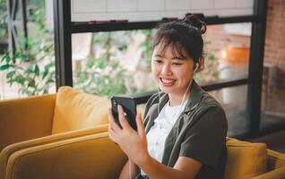 Smiling young asian woman using mobile phone checking social media,app playing game, shopping online, ordering delivery relax while sitting on a couch at cafe with laptop computer photo