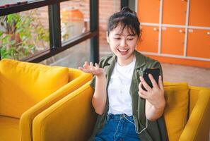 Smiling young asian woman using mobile phone checking social media,app playing game, shopping online, ordering delivery relax while sitting on a couch at cafe with laptop computer photo