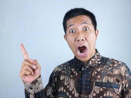 wow, surprised face expression asian man wearing batik clothes looking at camera while pointing side photo