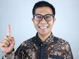 Smiling expression of asian man wearing batik clothes pointing up, promoting something photo