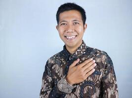 Smiling face expression of asian man wearing batik while hand on chest gesture photo