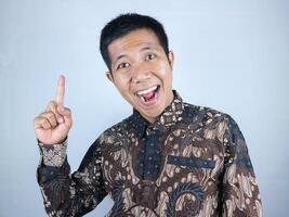 Smiling expression of asian man wearing batik clothes pointing up, promoting something photo