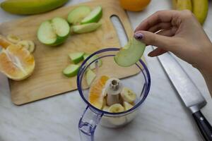 Girl throwing green apple slice into blender container while preparing sports cocktail in kitchen, ingredients for grinding in blender for healthy refreshing smoothie with vitamins photo