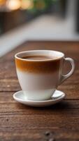 A white coffee cup with a brown foam on top sits on a wooden table. photo