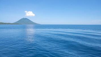 Volcanic Island in the Ocean. A volcanic island rises from the deep blue ocean, a single white cloud hangs above its peak. photo