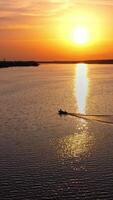 Aerial view over the water and motor boat floating at sunset. Beautiful view on the setting orange sun that reflects in water. Boat on the river moves to the industrial factory. Vertical video