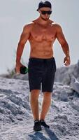 Handsome man with trained body holding flask in the mountains. Muscular athlete in shorts and sunglasses descends from the white hill in a sunny summer day. Vertical video