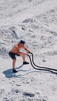 Strong young man doing exercises with a battle rope in sunny day. View from above on a muscular man training with cable on white ground outside. Concept of professional training. Vertical video