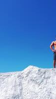 Drone view of a strong man in shorts without shirt on white hill. Young muscular man in good physical shape wearing sunglasses standing in nature. Camera moves up. Vertical video