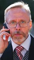Serious businessman wearing eyeglasses talks on phone while looking at camera. Portrait of a senior man in suit and tie talking on a cell phone in the city. Vertical video