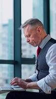 Thoughtful middle aged businessman in suit with a laptop sitting near the window working with documents, typing. Business . Vertical video