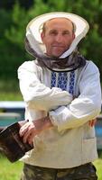 Beekeeper on apiary. Portrait of smiling man on outdoor hives background. Farmer apiarist in protective hat and in embroidery shirt looking on camera in summer day. Vertical video