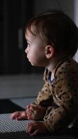 Lovely child in brown romper sitting at the wall indoors. Curious kid looks somewhere and then starts to crawl there. Side view. Vertical video