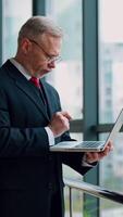 Mature senior serious businessman with laptop in hands thinking while checking e-mails standing near window with panoramic city view. from the side. Vertical video
